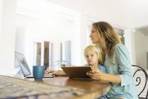 Garçon assis sur les genoux de sa mère et regardant une tablette pendant que sa mère travaille sur un ordinateur portable — Photo de stock