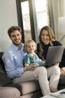 Retrato de padres felices e hijo sentado en el sofá con el ordenador portátil en casa - foto de stock