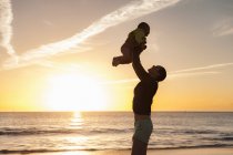 Mãe levantando a filhinha na praia ao pôr do sol — Fotografia de Stock