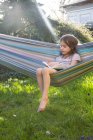 Little girl sitting on hammock in the garden reading a book — Stock Photo