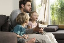 Father and children blowing into sails of toy model ship on couch at home — Stock Photo