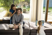 Couple with heaphones and daughter with laptop on sofa at home — Stock Photo