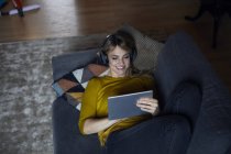 Портрет усміхненої жінки, що лежить на дивані і використовує навушники з планшетом — стокове фото