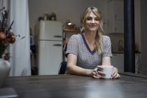 Retrato de mulher sorridente com xícara de café sentado à mesa na cozinha — Fotografia de Stock