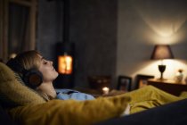 Mulher sorridente ouvindo música com fones de ouvido no sofá em casa à noite — Fotografia de Stock