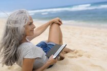 Bella donna anziana sorridente sdraiata sulla spiaggia, con in mano e-book — Foto stock