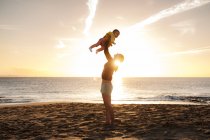 Mãe levantando a filhinha na praia ao pôr do sol — Fotografia de Stock