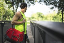 Runner standing on bridge in park, carrying bag and shoes — Stock Photo