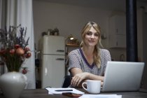 Portrait of smiling woman with laptop sitting at table in kitchen — Stock Photo