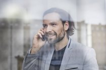 Retrato del hombre de negocios sonriente hablando de teléfono móvil detrás del cristal de la ventana - foto de stock