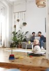 Los padres sentados en el sofá, utilizando el ordenador portátil, mientras que la hija está jugando en el suelo - foto de stock