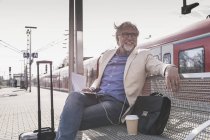 Empresário maduro sorridente sentado na estação de trem com telefone celular, fones de ouvido e caderno — Fotografia de Stock
