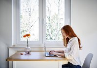 Rothaarige Frau sitzt mit Laptop am Tisch vor Fenster — Stockfoto