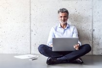 Retrato de hombre de negocios maduro seguro sentado en el suelo con el ordenador portátil - foto de stock
