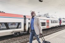 Mature businessman walking at train platform with earbuds and suitcase — Stock Photo