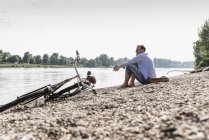 Uomo maturo con bici seduto sulla riva del Reno — Foto stock