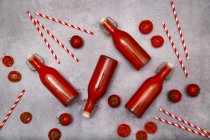 Jugo de tomate casero en botellas, pajitas y tomates batientes sobre tierra gris - foto de stock