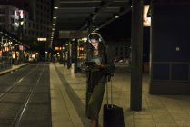Jovem com fones de ouvido e tablet esperando na estação à noite — Fotografia de Stock