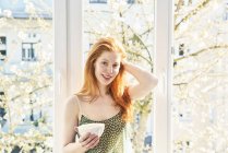 Portrait of redheaded woman with bowl of white coffee in front of window — Stock Photo