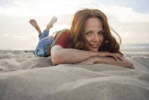 Redheaded woman lying in sand on the beach, smiling — Stock Photo