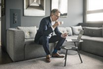 Reifer Geschäftsmann mit Tasse Kaffee und Laptop auf der Couch — Stockfoto