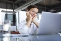 Усміхнена бізнесжінка сидить за скляною столиком в офісі дивлячись на ноутбук — стокове фото