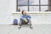 Junger Mann mit Skateboard sitzt auf dem Boden und benutzt Smartphone — Stockfoto