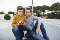Vater und Sohn sitzen auf Basketballplatz und nutzen Handy — Stockfoto