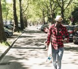 Joven y genial con monopatín caminando por la ciudad, usando un smartphone - foto de stock