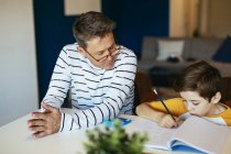 Pai assistindo filho fazendo lição de casa na mesa em casa — Fotografia de Stock