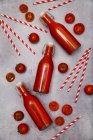 Jugo de tomate casero en botellas, pajitas y tomates batientes sobre tierra gris - foto de stock