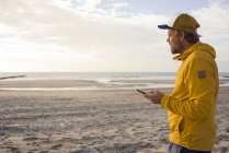 Человек в жёлтой куртке, со смартфоном на пляже — стоковое фото