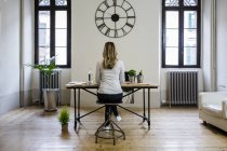 Rückansicht einer Frau, die zu Hause unter einer großen Wanduhr am Schreibtisch sitzt — Stockfoto