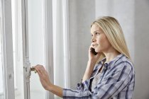 Mulher loira ao telefone olhando pela janela — Fotografia de Stock