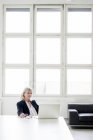 Улыбающаяся взрослая деловая женщина, работающая на ноутбуке в офисе — стоковое фото