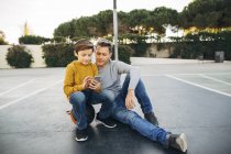Отец и сын сидят на баскетбольной площадке и пользуются мобильным телефоном — стоковое фото