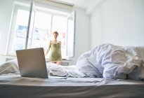 Laptop na cama com mulher de pé na frente da janela no fundo — Fotografia de Stock
