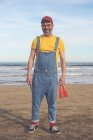 Ritratto di uomo sorridente in salopette in piedi sulla spiaggia con in mano bottiglie di bibite — Foto stock