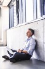 Mature businessman with laptop sitting on floor and leaning against concrete wall — Stock Photo