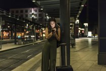 Jeune femme qui attend à la gare la nuit en utilisant un téléphone portable — Photo de stock