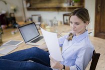 Femme assise au bureau à la maison les pieds levés et lisant le document — Photo de stock