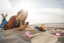Redheaded woman lying on the beach with beach toys, using smartphone — Stock Photo