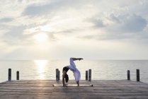 Giovane donna che pratica yoga su un molo vicino al mare al tramonto, urdhva dhanurasana — Foto stock