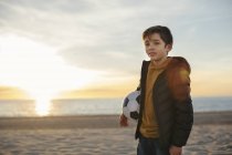 Retrato de niño sosteniendo pelota de fútbol en la playa al atardecer - foto de stock