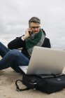 Ritratto di uomo d'affari al telefono seduto sulla spiaggia in inverno e utilizzando il computer portatile — Foto stock