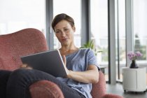 Mujer sentada en sillón en casa usando tableta - foto de stock