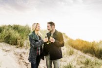 Happy couple in dunes clinking beer bottles — Stock Photo