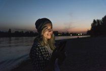 Jovem mulher usando smartphone à beira do rio à noite — Fotografia de Stock