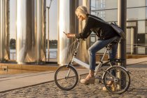 Mulher sênior com bicicleta da cidade usando telefone celular na beira do rio ao pôr do sol — Fotografia de Stock