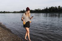Mujer joven caminando descalza a orillas del río, auriculares y teléfonos inteligentes - foto de stock